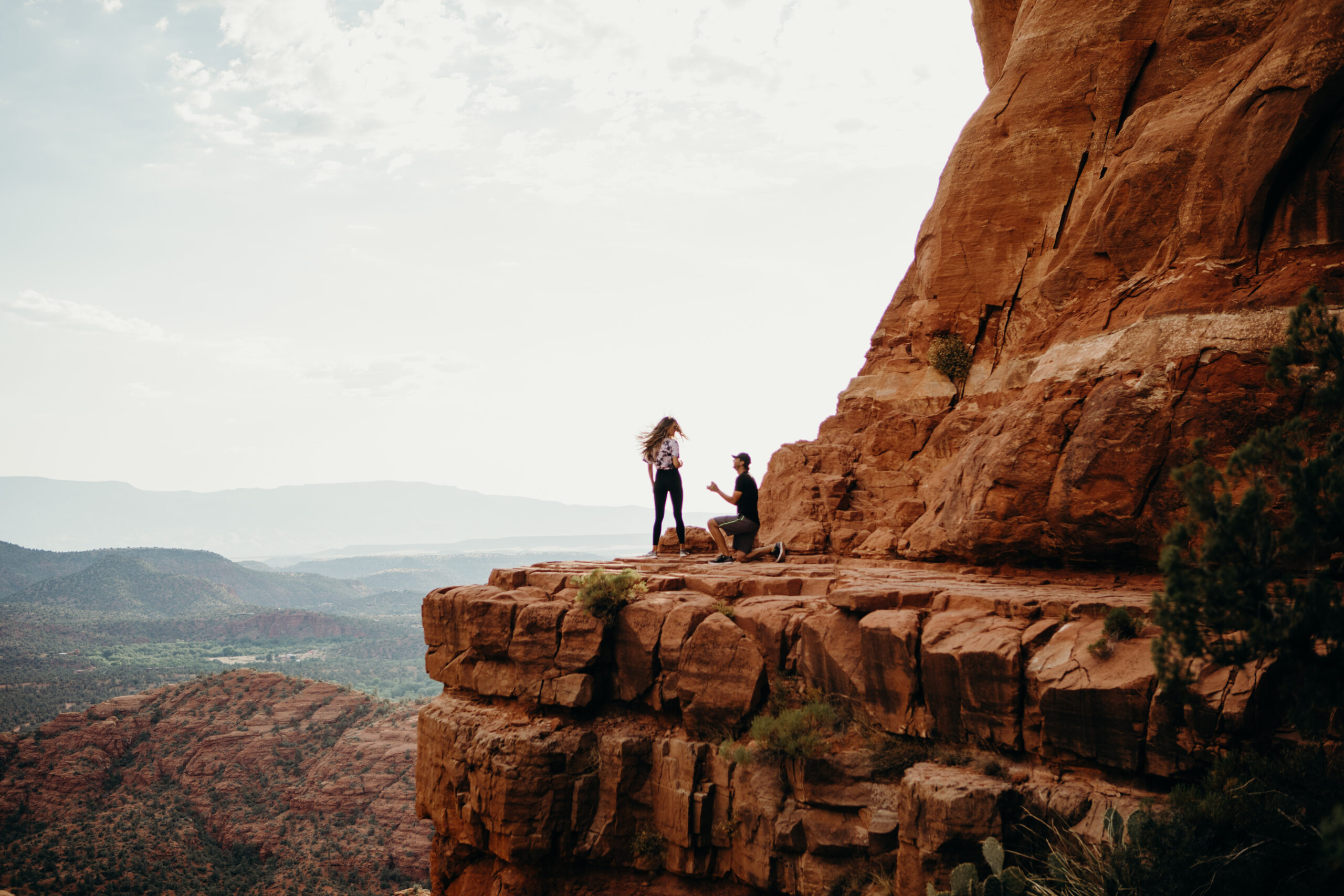 man is on one knee proposing to a woman on the ledge of cathedral rock