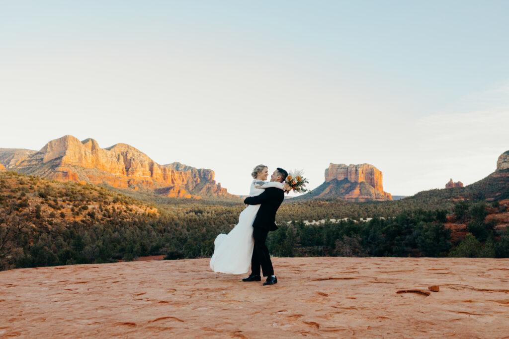 man holds woman in lace wedding dress on the base of cathedral rock swaying her