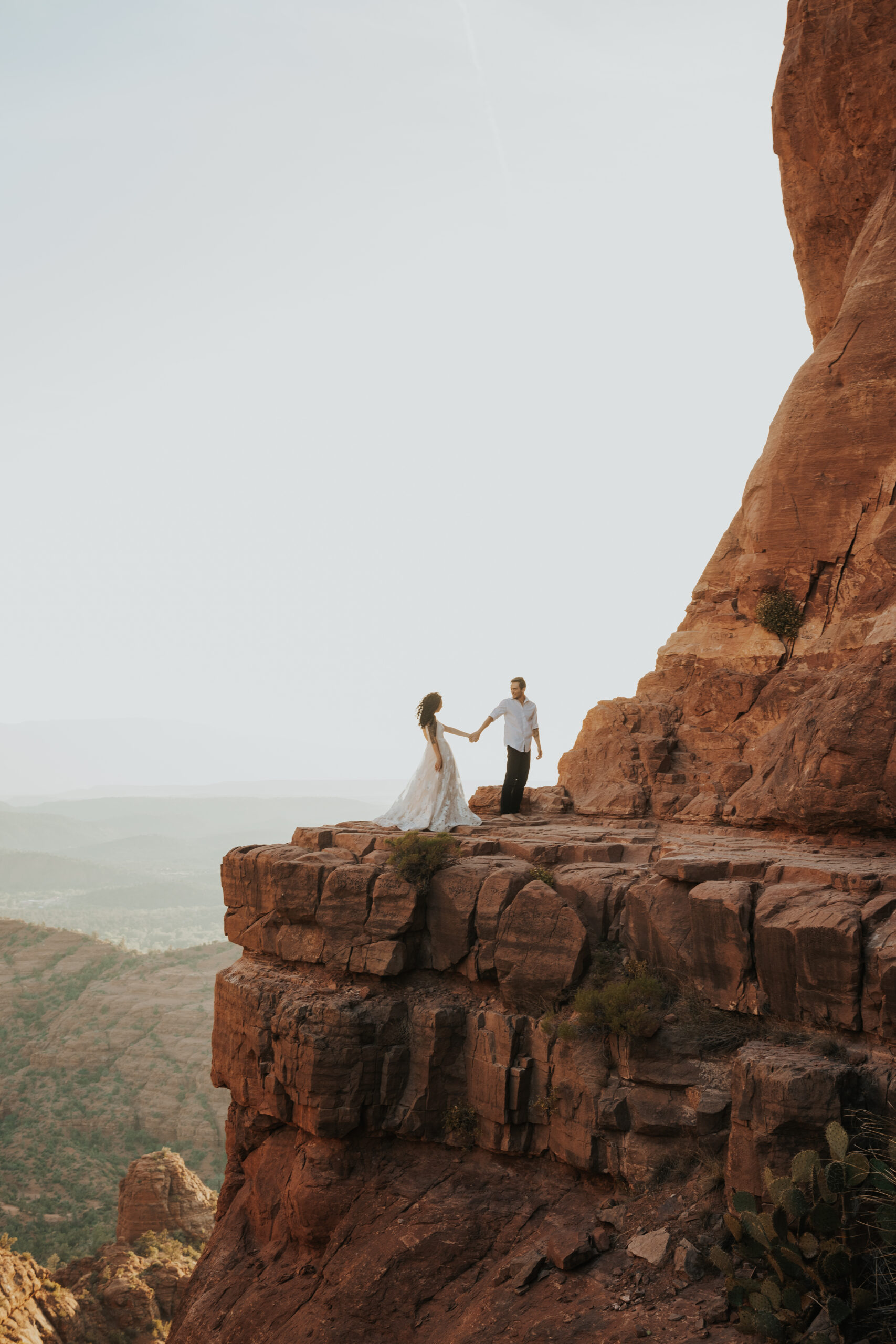 a man and woman in wedding dress and dress clothes hold hands on the ledge of cathedral rock