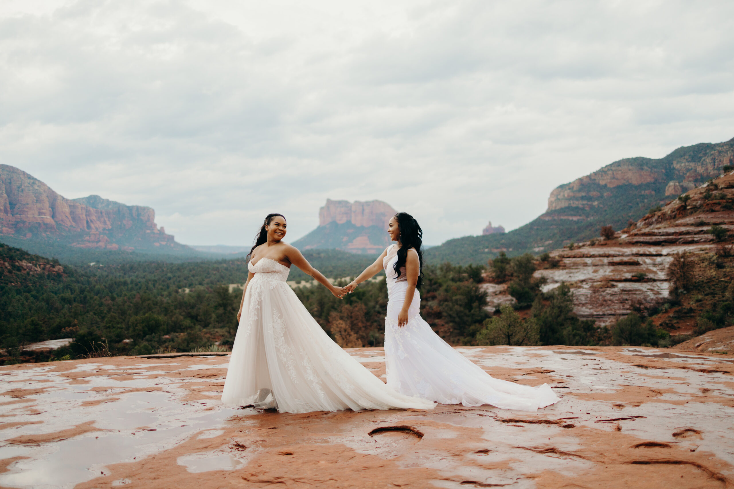 a woman holds her wife's hand on the base of cathedral rock both in wedding dresses amongst views of red rocks on their elopement day