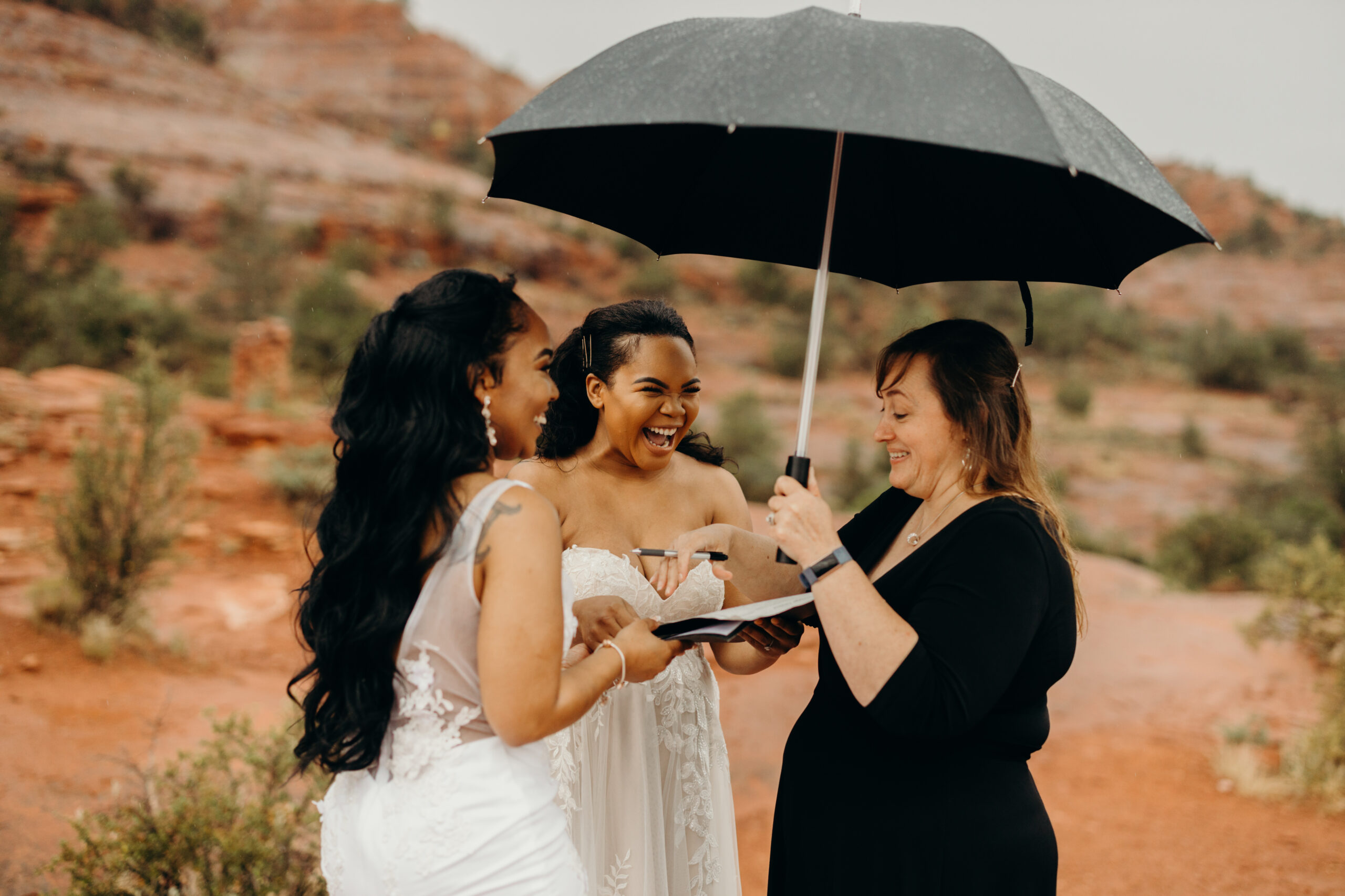 an officiant holds a black umbrella as she marries two woman in wedding dresses on cathedral rock